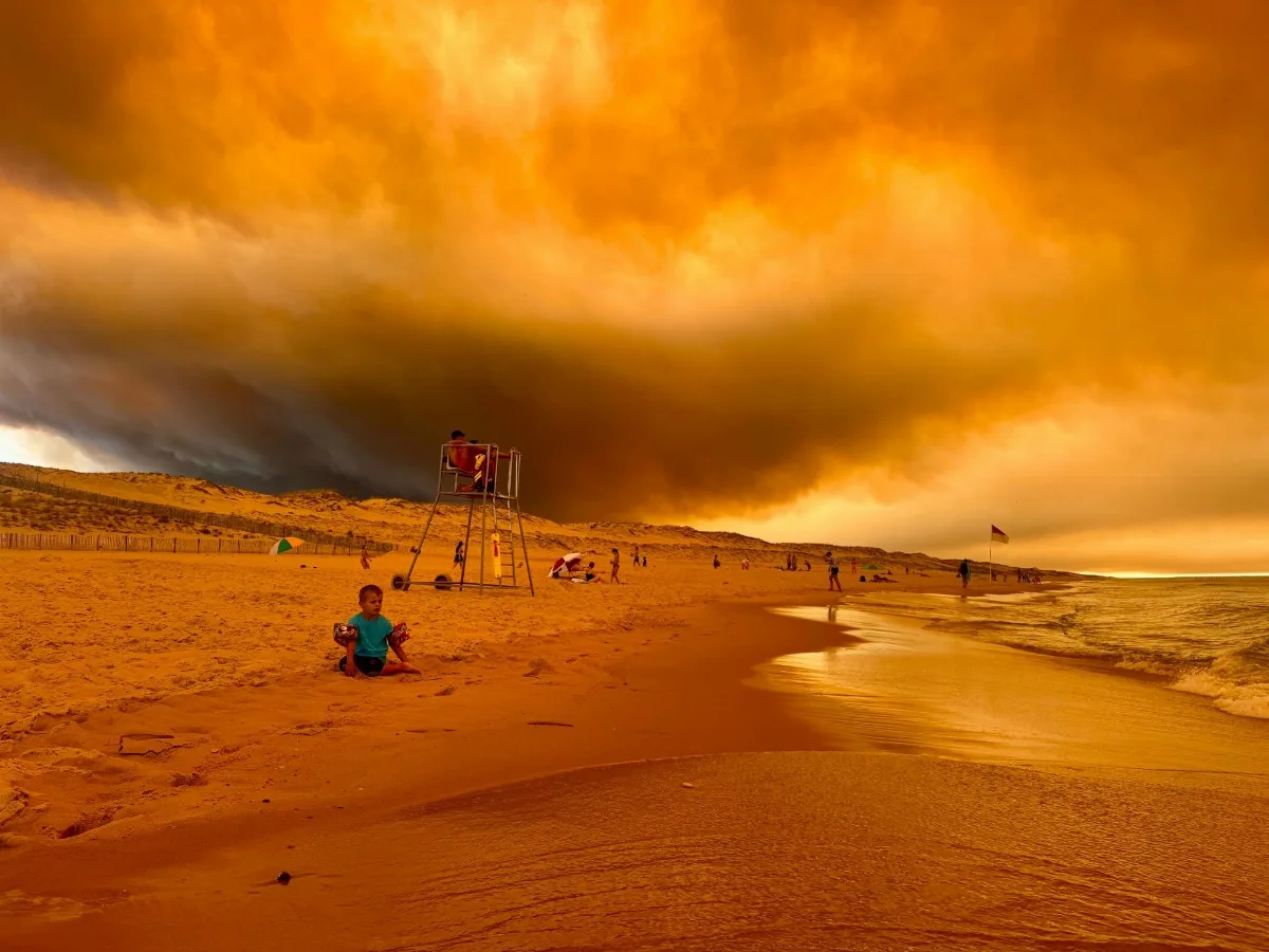 Ein kleines Kind mit blauem T-Shirt sitzt an einem Strand mit Weltuntergangsstimmung. Rechts ist das Meer. Im Hintergrund ist eine riesige Aschewolke zu sehen. Das gesamte Bild ist orange-rötlich durch die Asche in der Luft.
Es sind mehr Menschen in der Ferne am Strand zu sehen.