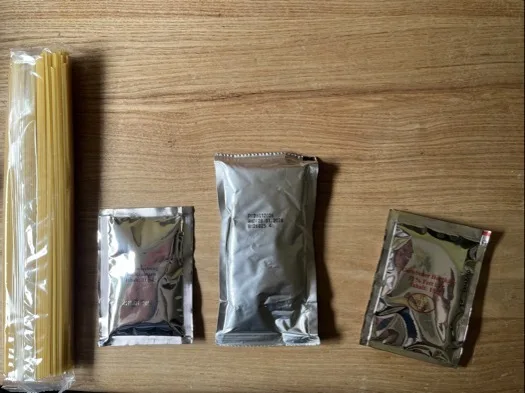 Auf einer hellen Holzarbeitsfläche liegt links ein Bündel ungekochter Spaghetti in durchsichtiger Plastikfolie. Rechts davon in einer Reihe drei silberne Folienpackungen in unterschiedlicher Größe, die rechte Packung zeigt teilweise einen aufgedruckten bunten Aufdruck mit roten Schriftzeichen.