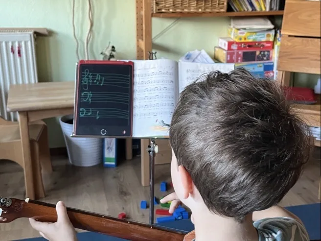Ein Kind von hinten sitzt mit einer Gitarre und hält das Griffbrett mit der linken Hand. Vor ihm steht ein Notenpult mit einer aufgeschlagenen Notenheftseite rechts und einem Tablet oder Schreibtafel mit grüner handschriftlicher Notation links; im Hintergrund Regale, Brettspiele, Holzmöbel und Spielzeugwürfel auf dem Boden.