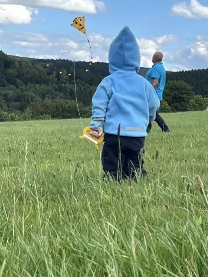 Ein kleines Kind von hinten mit hellblauer Kapuzenjacke hält in der rechten Hand eine gelbe Griffrolle, an der eine dünne Schnur nach oben zu einem gelben Drachengesicht-Kite verläuft. Im Hintergrund steht ein Erwachsener in türkisblauem Oberteil auf einer großen, grasbewachsenen Wiese vor einem Baumsaum und einem bewölkten Himmel.
